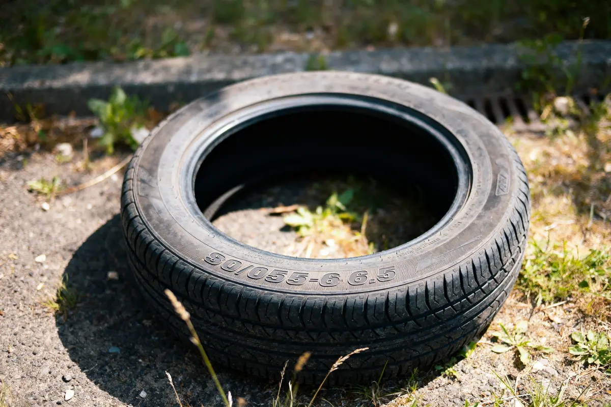 Pneu noir usé, abandonné sur une route en asphalte, entouré de végétation et bordure de trottoir.