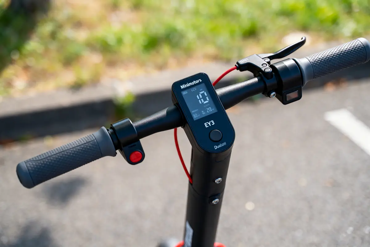 Guidon de trottinette électrique avec écran numérique, manettes noires et câblage rouge. Route floue en arrière-plan.