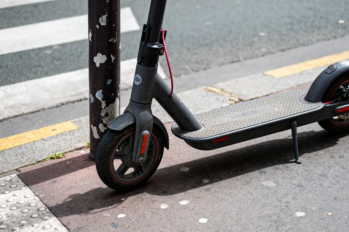 Trottinette électrique grise garée sur trottoir près passage piéton, attachée à un poteau noir et blanc.