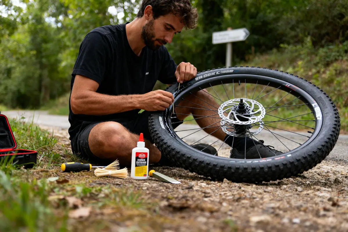 Un homme répare une roue de vélo sur un chemin forestier avec des outils à proximité.