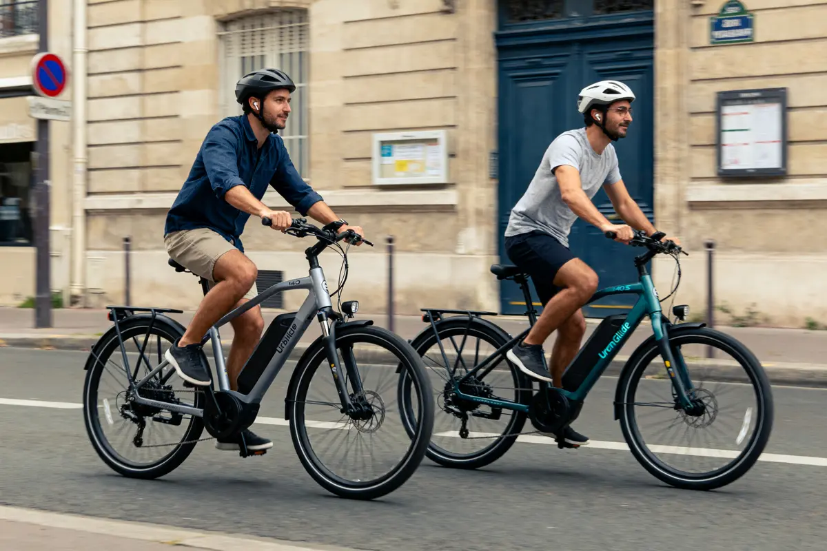 Deux hommes à vélo électrique, portant des casques, circulent en ville devant un bâtiment en pierre.