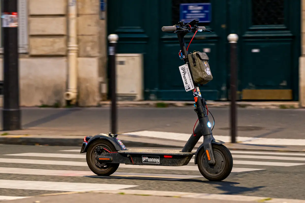 Trottinette électrique noire sur passage piéton urbain, sac accroché au guidon, environnement urbain en arrière-plan.